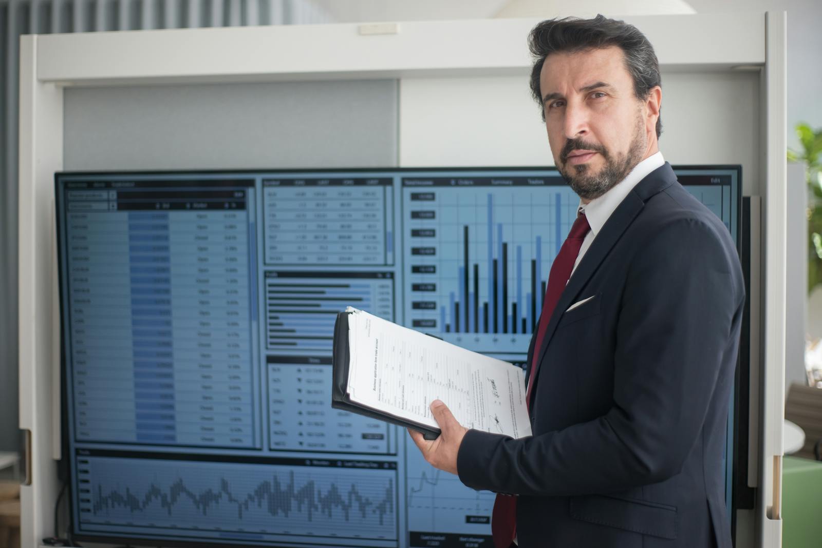 Businessman in a suit holds documents analyzing graphs on a screen, reflecting data analysis.